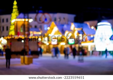 Blurred background. People walk in city square on winter night. Black silhouettes of people walking near houses decorated luminous illumination. White light bokeh blur spots from glowing house lights