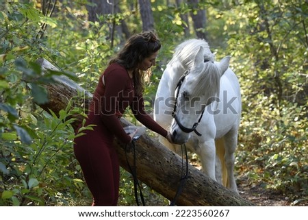 A girl gives her white horse something to eat before jumping over a tree. Scene in the forest with the teacher and her white horse. The white horse eats from the girl's hand.