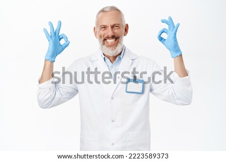 Smiling senior doctor, medical worker showing okay sign, recommending clinic, standing over white background.
