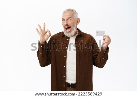 Smiling senior man showing credit card and okay sign, recommending bank, winking at camera, standing over white background.