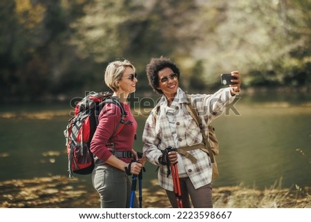 Two female friends taking selfies by a lake during a hike along the mountain.