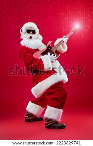 Studio portrait of Santa Claus in sunglasses and headphones playing the guitar and singing. Merry Christmas. Party. Full length shot. Festive red background with lights. 