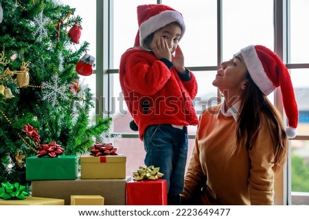 Happy Asian family mom and daughter in red and white Santa Claus hat and sweater smiling playing together near Christmas pine tree with glitter decorating items and present gift boxes in xmas evening.