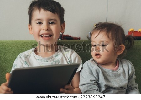 Brother relaxing on sofa in living room with her little baby sister while watching cartoon series online. Cute boy and girl using digital tablet at home. Happy Royalty-Free Stock Photo #2223665247