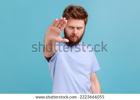 Portrait of strict bearded man showing stop sign at camera, standing with frowning face and bossy expression, meaning caution to avoid danger or mistake. Indoor studio shot isolated on blue background
