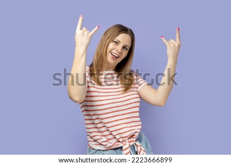 Yeah, awesome. Excited rocker blond woman wearing striped T-shirt showing rock and roll hand gesture, crazy devil horns with fingers and yelling. Indoor studio shot isolated on purple background.