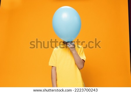 a boy of school age stands on an orange background in a yellow polo and covers his face with a large blue balloon