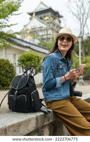 vertical shot fashionable Asian girl wearing hat and sunglasses sitting relaxing on a step and holding phone with smile on face while visiting Osaka castle in japan