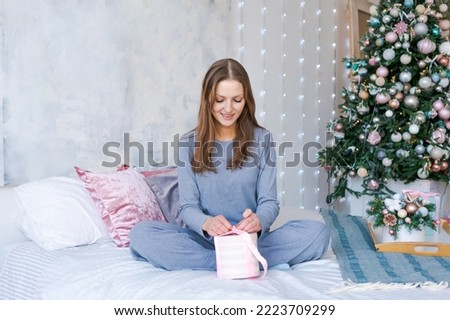 New Year's photo girl on bed. Portrait young woman in blue pajamas, without makeup in a room against the background of a Christmas tree, sits happy with a gift box in her hand