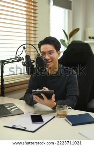 Smiling asian male podcaster sitting front of condenser microphone and at home studio and smiling to camera