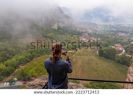 Tourist woman taking pictures from Holy Monastery of Rousanou in Kalambaka, Meteora, Thessaly, Greece, Europe. Unique rock formations are surrounded by mystical fog. Moody dramatic atmosphere