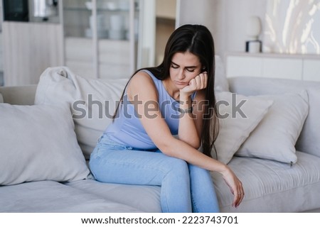 Disappointed young beautiful Hispanic girl sitting on cozy sofa with cushions leans on hand looks down in upset mood. Troubles, financial problems. Perplexed female at home. Failure, lack of energy. Royalty-Free Stock Photo #2223743701