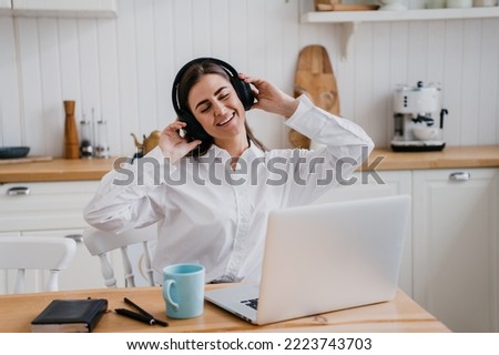 Cheerful brunette young woman in white shirt sitting at table with laptop eyes closed listening music using headphones. Pretty hispanic girl at break of remote learning. Domestic leisure and music