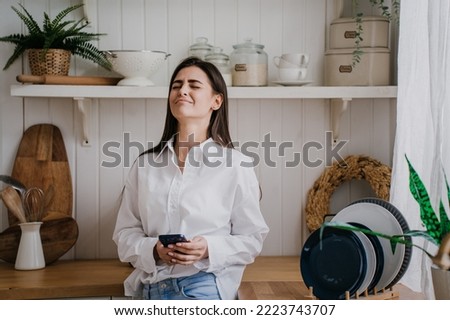 Upset brunette young woman in white shirt crying eyes closed with frustrated face expression holds phone standing at kitchen. Grief, troubles, failure. Pretty Hispanic girl after divorce.