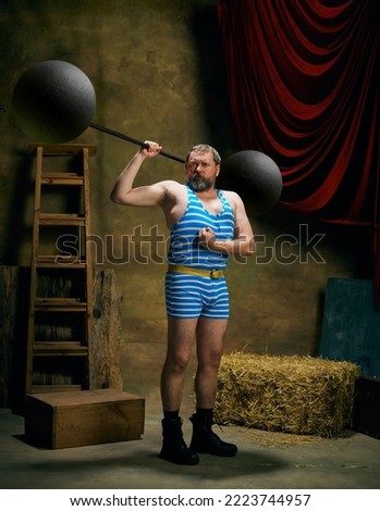 Retro circus. Cinematic portrait of retro circus strongman wearing striped sports swimsuit holding barbell on dark circus backstage background. Concept of creative art, fashion, style and inspiration Royalty-Free Stock Photo #2223744957