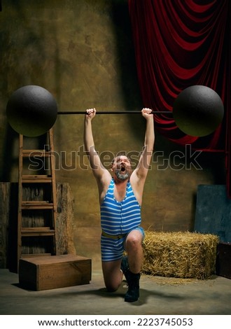 Circus athlete. Vintage portrait of retro circus strongman wearing blue striped sports swimsuit training with barbell over dark circus backstage background. Concept of creative art, fashion, style