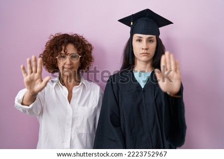 Hispanic mother and daughter wearing graduation cap and ceremony robe doing stop sing with palm of the hand. warning expression with negative and serious gesture on the face. 