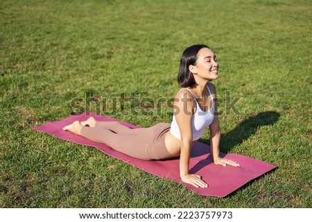 Smiling young sportswoman stretches on rubber mat in park, does yoga asana exercises, workout on fresh air in fitness clothing.