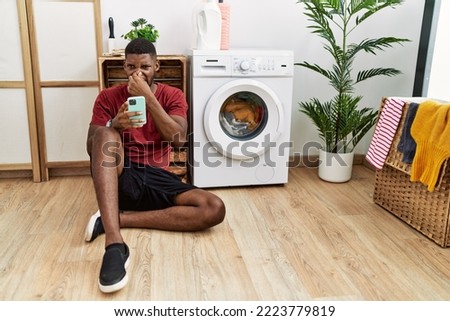 Young african american man using smartphone waiting for washing machine smelling something stinky and disgusting, intolerable smell, holding breath with fingers on nose. bad smell 