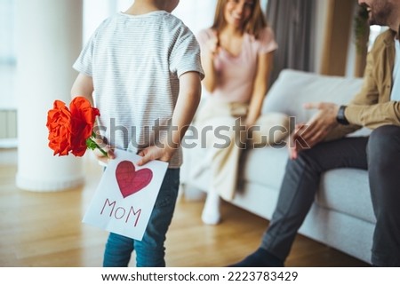 Happy little boy congratulating smiling mother and giving card with red heart during holiday celebration at home. Little boy giving card to mom. Mother and son reading greeting card 