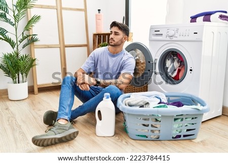 Young hispanic man putting dirty laundry into washing machine looking to side, relax profile pose with natural face and confident smile. 