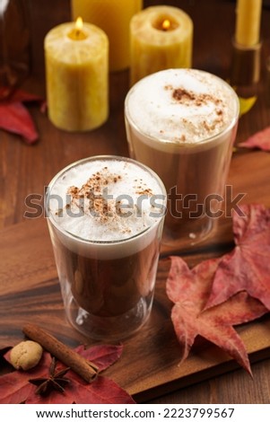 Two transparent glasses with cappuccino with black coffee, milk foam and cinnamon in autumn setting, burning candles, pumpkins, yellow and red tree leaves on wooden background