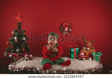 photo of a nice boy dressed as an elf next to a Christmas decoration with a tree and gifts