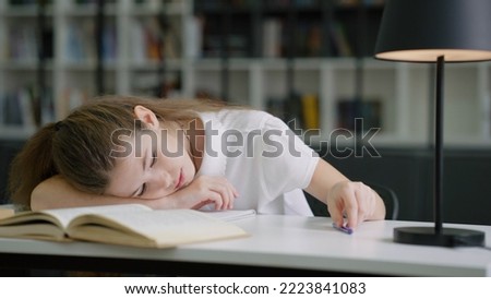 Tired school girl lying on desk and playing with pen to entertain herself, pupil at boring lesson at school, bookshelves on blurred background. Concept of education
