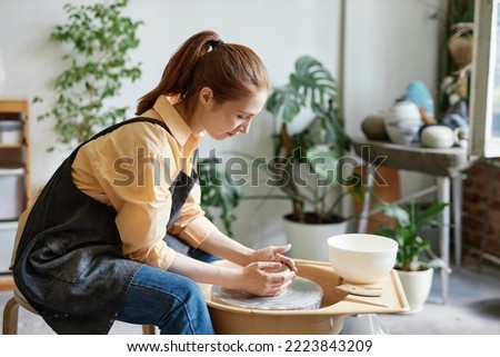 Side view portrait of young woman using pottery wheel and creating handmade ceramics in cozy studio