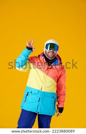 smiling man in goggles and snowboard clothes doing an ok with raised hand on a yellow background.