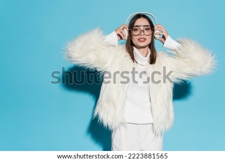 stylish young woman in eyeglasses and faux fur jacket holding winter earmuffs on blue Royalty-Free Stock Photo #2223881565