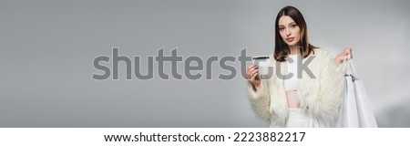 Young woman in faux fur jacket holding white shopping bags and credit card on grey background, banner