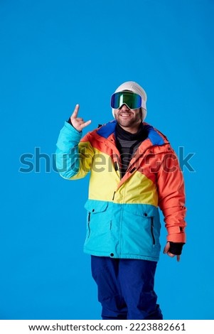 Man in snowboarding clothes and goggles smiling at the camera and making the horns with the fingers of his hand on a blue background.