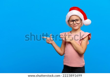 Happy new year concept. Smiling caucasian girl wearing glasses and santa claus red hat points at copy space on blue isolated background