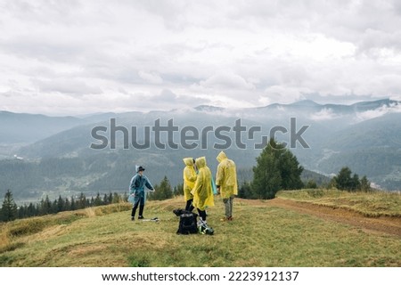 A group of young tourists in raincoats are standing in the mountains against the background of a beautiful view during a stopover and are about to continue their journey.
