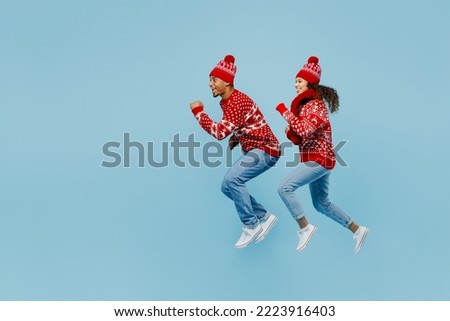 Full body side view merry fun young couple two man woman wear red Christmas sweater Santa hat posing jump high run isolated on plain pastel light blue background. Happy New Year 2023 holiday concept