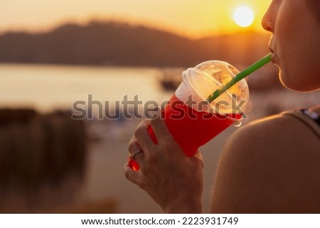 Unrecognizable young woman in the foreground enjoying a slushy at sunset while drinking through a straw. Royalty-Free Stock Photo #2223931749