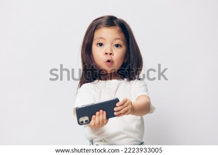  little girl in a white t-shirt stands on a light background with a fashionable smartphone in her hand and takes a selfie, looking at the phone. photography with empty space for advertising mockup