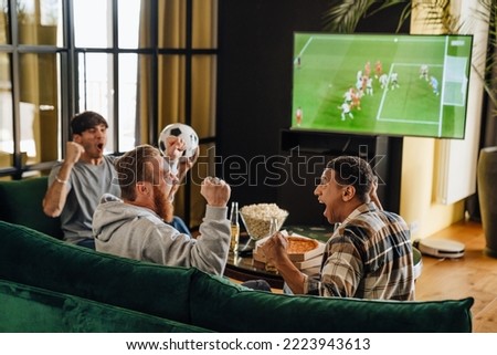 Male friends gesturing as winners while watching football match sitting on sofa in front of the TV screen Royalty-Free Stock Photo #2223943613