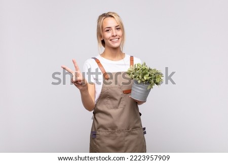 young woman gardener smiling and looking friendly, showing number two or second with hand forward, counting down