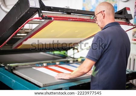 In the printing house, an experienced technician works on a screen printing machine. Production work. Check the print quality.