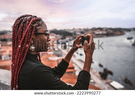 A profile shot of a young African female traveler with red box braids shooting a tourist attractions with her smartphone; a black tourist woman photographing urban landscape panorama via the cellphone