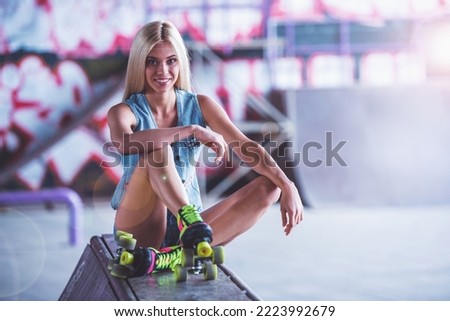 Beautiful blonde girl in rollers is looking at camera and smiling while sitting in skatepark