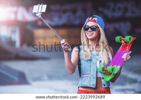 Beautiful blonde girl in cap and glasses is making selfie using a smart phone and smiling while standing with her skateboard in skate park