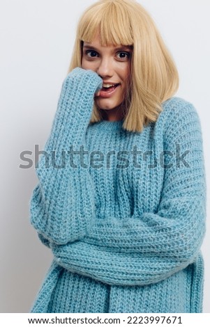 vertical photo of a beautiful, pleasant blonde woman in a long blue sweater, smiling broadly at the camera, holding her hand near her face