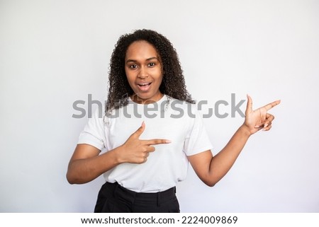 Portrait of amazed young woman pointing aside with fingers over white background. African American woman wearing white T-shirt advertising something. Promotion concept