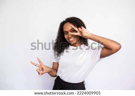 Portrait of cheerful young woman peeking through fingers. African American lady wearing white T-shirt making peace gesture and smiling over white background. Fun and happiness concept