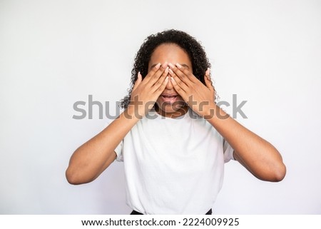Portrait of curious young woman covering eyes with hands over white background. African American lady wearing white T-shirt anticipating surprise or playing peekaboo. Surprise concept
