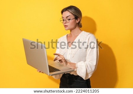 Young nerdy woman wearing eyeglasses is using laptop computer on yellow background