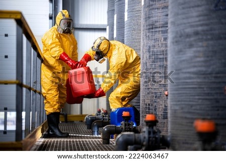 Chemical worker carrying canisters with hazardous materials and standing by large storage acid tanks. Royalty-Free Stock Photo #2224063467